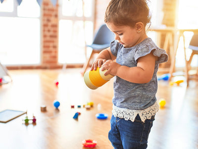 A toddler holding a yellow cup in a sunlit room with scattered toys
