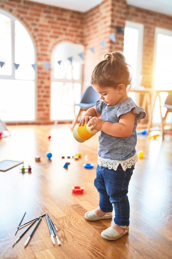 A toddler holding a yellow cup in a sunlit room with scattered toys